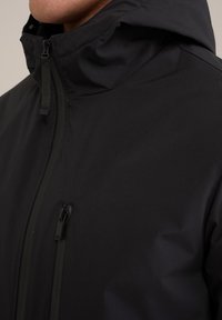 Chaqueta negra con cremallera, cuello alto y capucha. Presenta una textura suave, acabado mate y costuras detalladas. Diseño compacto ideal para superposiciones.