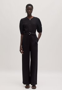 Zwarte jumpsuit met korte pofmouwen, rits aan de voorkant en riem in de taille. Heeft brede pijpen en subtiele zakdetails. Gladde fabric textuur.
