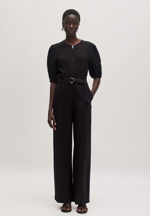 Combinaison noire avec des manches courtes bouffantes, fermeture éclair sur le devant et taille ceinturée. Présente des jambes larges et des détails de poches subtils. Tissu au texture lisse.