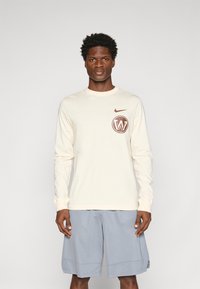 Kermanen pitkähihaine paita, jossa on ruskea logo rintataskussa, yhdistettynä sinisiin shortseihin. Pehmeä rakenne, rento muotoilu, pyöreä pääntie ja pitkät hihat.