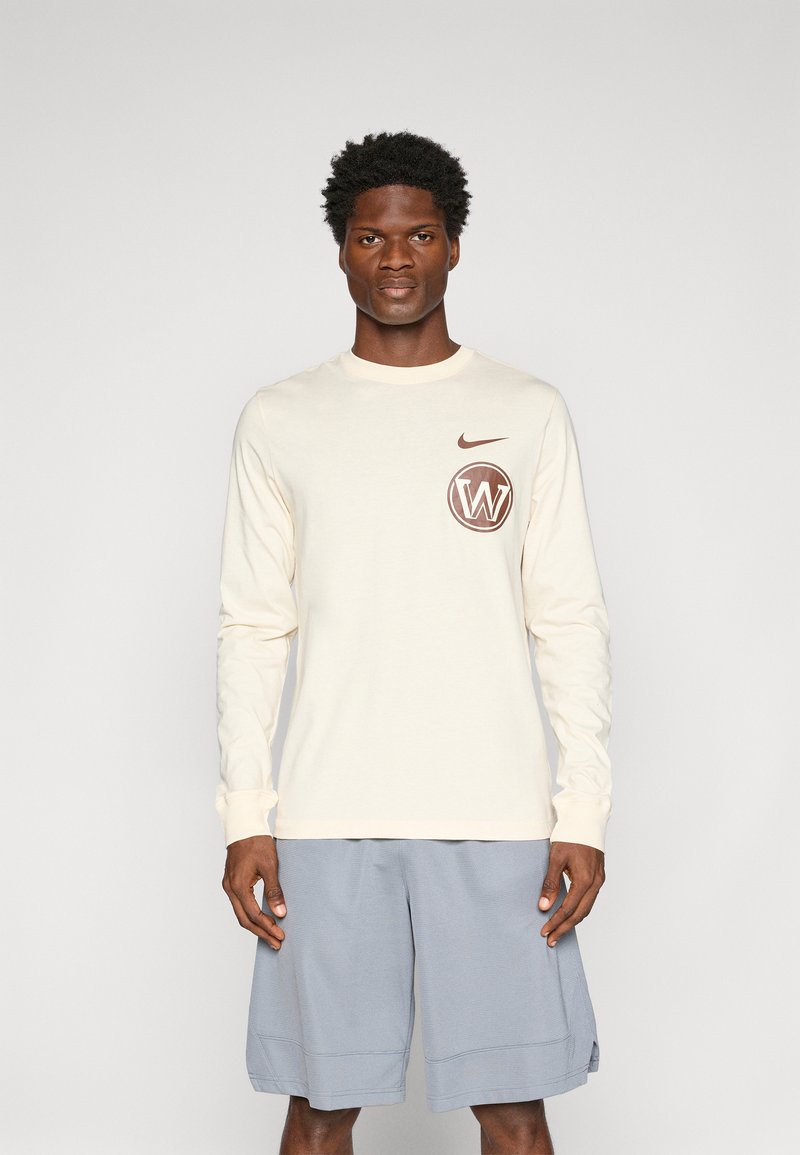 Kermanen pitkähihaine paita, jossa on ruskea logo rintataskussa, yhdistettynä sinisiin shortseihin. Pehmeä rakenne, rento muotoilu, pyöreä pääntie ja pitkät hihat.