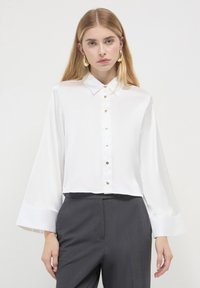 Chemise blanche en satin à manches longues et larges, avec des boutons dorés sur le devant, associée à un pantalon taille haute gris foncé.