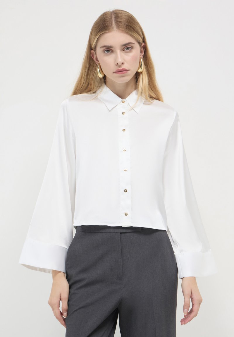 Chemise blanche en satin à manches longues et larges, avec des boutons dorés sur le devant, associée à un pantalon taille haute gris foncé.