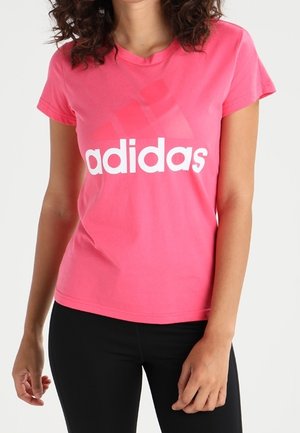 Kobieta ubrana w jaskraworóżowy t-shirt Adidas z białym logo i czarne spodnie, stojąca na jednolitym jasnym tle.
