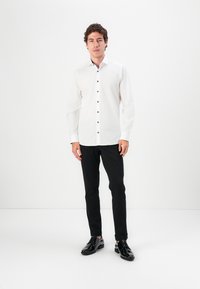 Camisa blanca de manga larga con botones negros, combinada con pantalones negros y zapatos negros brillantes, sobre un fondo blanco liso.