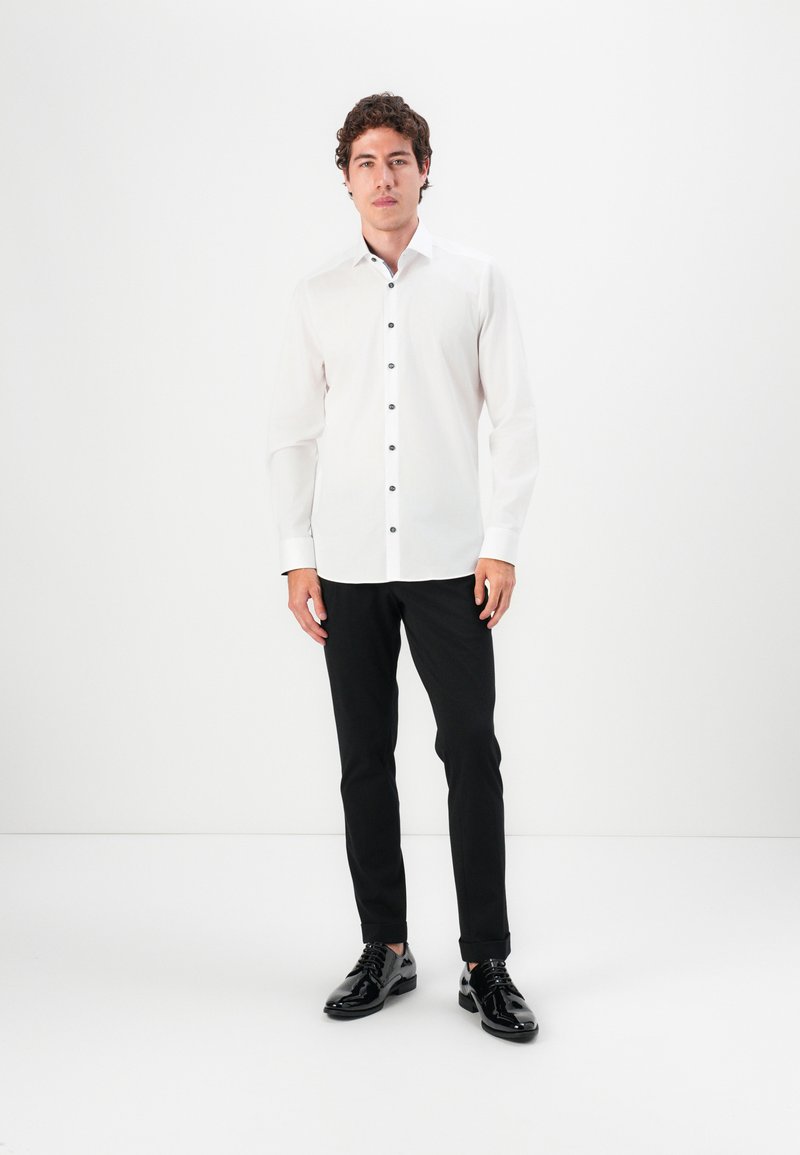 Camisa blanca de manga larga con botones negros, combinada con pantalones negros y zapatos negros brillantes, sobre un fondo blanco liso.