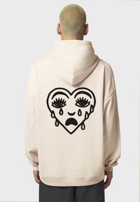 Felpa beige con un grande cuore ricamato nero che presenta occhi in lacrime e lacrime, realizzata in tessuto morbido con una vestibilità comoda.