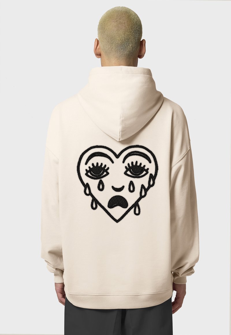 Felpa beige con un grande cuore ricamato nero che presenta occhi in lacrime e lacrime, realizzata in tessuto morbido con una vestibilità comoda.