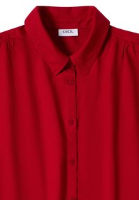 Blouse rouge boutonnée avec col pointu et détail de tissu froncé près des coutures d’épaule, étiquetée « CECIL » à l’intérieur du col.