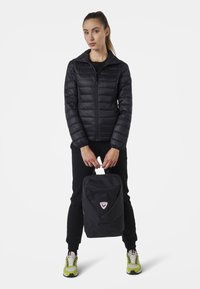 Giacca a piumino nera con chiusura a zip, abbinata a uno zaino nero con un logo triangolare e una tasca con zip. Il modello indossa pantaloni neri e sneakers.