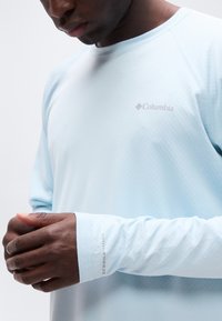 Άνδρας που φοράει μακρυμάνικο μπλε ανοιχτό πουκάμισο Columbia με το κείμενο "Omni-Freeze" στο μανίκι, ρυθμίζοντας τη μανσέτα του με τα δύο χέρια.