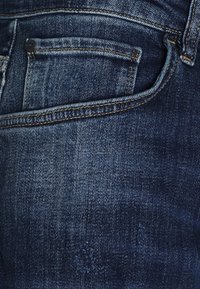 Blue denim jeans featuring a back pocket with stitching details and a slight worn texture, emphasizing a medium wash finish.