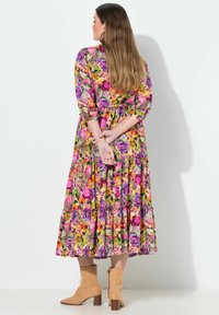 Květinové maxi šaty s živými vzory v barvách fialové, růžové a oranžové. Mají tříčtvrteční rukávy, vrstvenou sukni a pásek. Světle hnědé kotníkové boty.