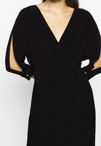 El vestido negro de tejido suave presenta un escote en V profundo, mangas fluidas con aberturas y un cinturón a juego. Acentuado con botones decorativos en las mangas.