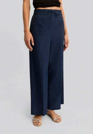 Donna che indossa pantaloni a gamba larga blu scuro con vita elasticizzata e coulisse, abbinati a sandali beige con cinturini e top corto nero.