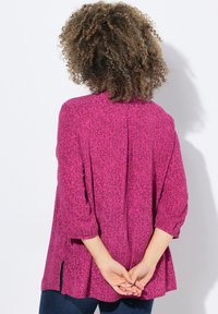 Blusa magenta con motivo floreale, taglio morbido, maniche a tre quarti e spacchi laterali. Il materiale sembra leggero e texturizzato.