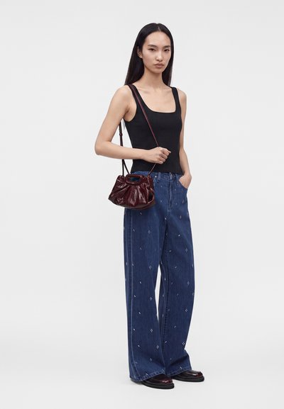 Femme portant un débardeur noir, un jean bleu à jambes larges avec de petits motifs blancs, des chaussures noires, et un petit sac à bandoulière rouge foncé.