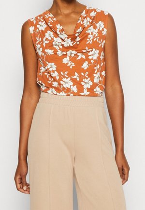 Ärmelloses orangenes Oberteil mit weißem Blumenmuster, kombiniert mit hochtaillierten beigen Hosen mit weitem Bein.