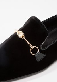 Mocassin en velours noir avec un accent métallique doré orné d'un design en forme de serrure et des détails en cuir, texture lisse et forme classique.