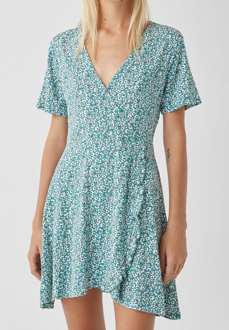 Robe à manches courtes, couleur turquoise avec un motif floral blanc, décolleté en V, design croisé, ourlet volanté, tissu doux et coupe décontractée.