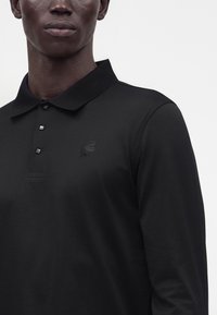 Homme portant un polo noir à manches longues avec un col texturé et un petit logo brodé sur la poitrine.