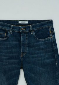 Jeans de mezclilla oscuros y ajustados con cierre de botón, presillas para cinturón, bolsillos delanteros y etiqueta de la marca "Salsa Jeans" en la cinturilla.