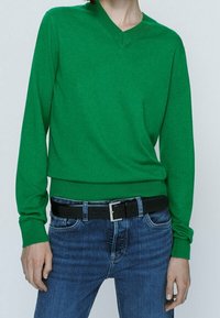 Vihreä V-päällysetakkikosteuttaja, joka on valmistettu pehmeästä neuleesta, pitkähihaiset hihat ja tiukasti istuva vyötärö, yhdessä sinisten farkkujen ja mustan vyön kanssa.