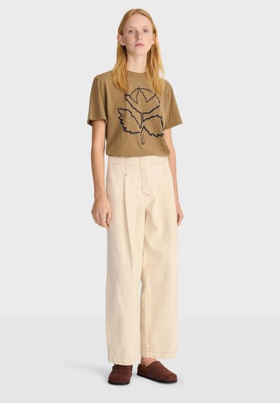 Jeune personne aux longs cheveux blonds portant un t-shirt marron à motif feuilles, un pantalon large couleur crème, des chaussettes rayées et des chaussures marron sans lacets.