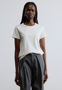 Jasnobeżowa koszulka z krótkim rękawem i okrągłym dekoltem, z małym jasnym logo na klatce piersiowej. Noszona z szarymi plisowanymi spodniami.