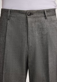 Pantaloni grigi a motivo chevron con bottoni e passanti per cintura, indossati con una camicia bianca infilata in vita.