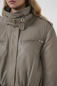 Bézs bőrdzseki magas gallérral, cipzáras mellzsebekkel és öves nyakdísszel. Simább textúra és strukturált kialakítás.