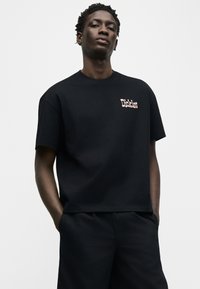 Νεαρός άνδρας με κοντά σγουρά μαλλιά, φορώντας μαύρο μπλουζάκι Dickies και μαύρο παντελόνι, στέκεται με τα χέρια στις τσέπες μπροστά σε απλό φόντο.