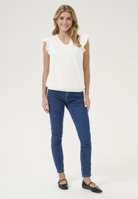 Haut blanc à col en V avec des manches courtes volantes, associé à un jean skinny bleu et des ballerines noires, se tenant sur un fond clair uni.