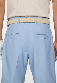 Ljusblå byxor i mjukt tyg med ett flätat beige bälte. Har två bakfickor och en avslappnad passform. Texturerad bälte detalj synlig.