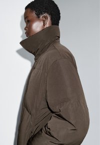 Brun pufferjakke med høj krave, lynlås og struktureret stof. Har en løs pasform og en subtil glans på overfladen.