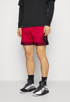 Person trägt ein schwarzes langärmeliges Oberteil, rote Basketballshorts mit schwarzen Streifen, schwarze Socken und schwarz-weiße Sportschuhe mit Logos.