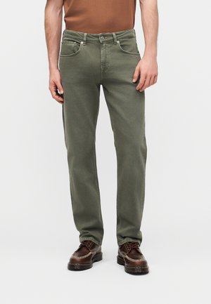 Man draagt olijfgroene jeans met rechte pijpen, bruine schoenen en een bruine, in de broek gestoken overhemd, staand tegen een effen lichte achtergrond.