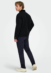 Črna kordurna jakna s ovratnikom, dolgimi rokavi in manšetami na gumbe, kombinirana z mornarsko modrimi hlačami in svetlimi športnimi čevlji.