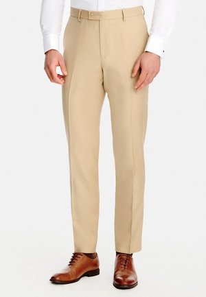 Mężczyzna ubrany w beżowe eleganckie spodnie, białą koszulę i brązowe skórzane eleganckie buty, stojący na tle jednolitego białego tła.