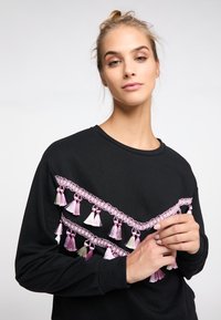 Hanorac negru cu ciucuri decorativi roz și alb aranjati într-un model în zigzag pe partea din față. Mâneci lungi cu manșete ribbate.