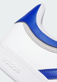 Zapatilla blanca con suela de goma texturizada. Presenta una superposición de cuero azul, acentos grises y malla blanca en el cuello. Logotipo de Adidas visible.