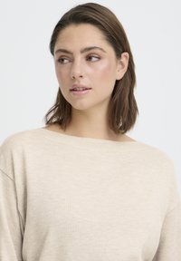 Bézs, bő szabású pulóver széles csónaknyakú kivágással és bordázott textúrával. Puha anyag, ejtett vállvarratokkal, minimalista dizájn.