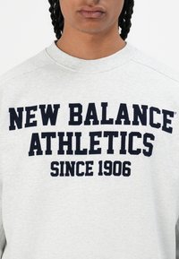 Ljusgrå sweatshirt med rund halsringning, med fet marinblå text: "NEW BALANCE ATHLETICS SINCE 1906" som framhävs.
