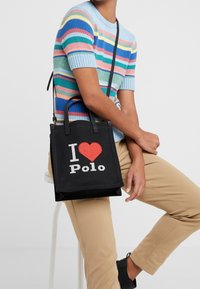 Černá plátěná taška s designem "I ❤️ Polo" v bílé a červené barvě. Krátké uši a nastavitelný ramenní popruh. Hladká textura.