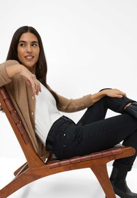 Cardigan lavorato a maglia beige, camicia bianca, jeans neri, seduto su una sedia di legno. Texture morbida, vestibilità rilassata, dettaglio dei bottoni sul cardigan.