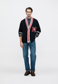 Μπλε σκούρα μπλε ζακέτα με πλέξη καλωδίου, με κόκκινο και λευκό περίγραμμα, κούμπωμα με κουμπιά και πατς στο στήθος, φορεμένη πάνω από καρό πουκάμισο και μπλε τζιν.