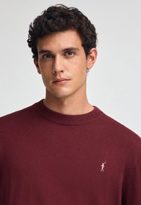 Bordowy sweter z ribbowanym okrągłym dekoltem, wykonany z miękkiej dzianiny, z małym białym emblematem na lewej piersi.