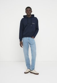 Μπλε ναυτικό βαμβακερό φούτερ με τσέπη μπροστά και κουκούλα με κορδόνι, σε συνδυασμό με ανοιχτό μπλε τζιν και μπεζ αθλητικά παπούτσια σε λευκό φόντο.