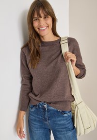 Brązowy sweter dzianinowy z okrągłym dekoltem, zestawiony z niebieskimi dżinsami i jasnokolorową torebką na ramię z paskiem.