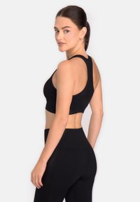 Jauna sieviete ar tumšiem matiem, sasietiem atpakaļ, ģērbusies melnā sporta krūšturī un augstām legingām, skatās pāri plecam uz baltā fona.